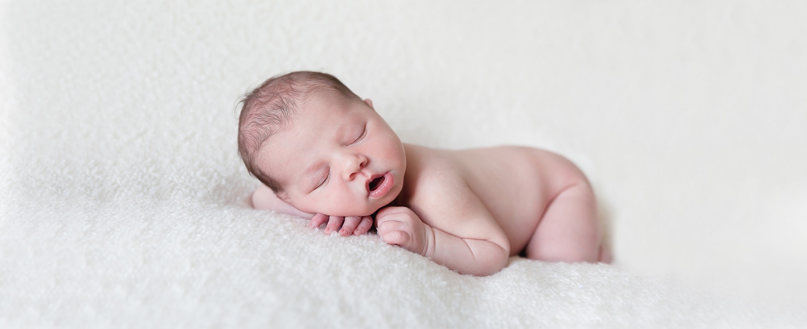 Il y a un bébé endormi posé sur le ventre la tete posée sur une de ses mains sur un plaid de couleur beige.